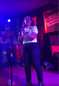 Caterina Ciananni, Executive Director of the United Workers Union, standing on stage and speaking into a microphone during her speech at the Semaphore Workers Club's at Bread and Roses event. She is wearing glasses, a white t-shirt with various prints, and dark jeans. A drum set is partially visible in the background, along with other decorative items.
