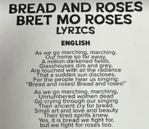 The image displays the lyrics of "BREAD AND ROSES" prominently in bold, capitalized letters at the top. Beneath the title, the word "ENGLISH" specifies the language of the following lyrics. The lyrics read: “As we go marching, marching, our home so far away, a million darkened fields, glasshouses dim and grey, are touched with all the radiance that a sudden sun discloses, for the people hear us singing: Bread and roses! Bread and roses!”