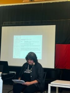 Associate Professor Natalie Harkin lectured at the event. She is seated and looking at a paper. In the background, there is a slide presentation. 