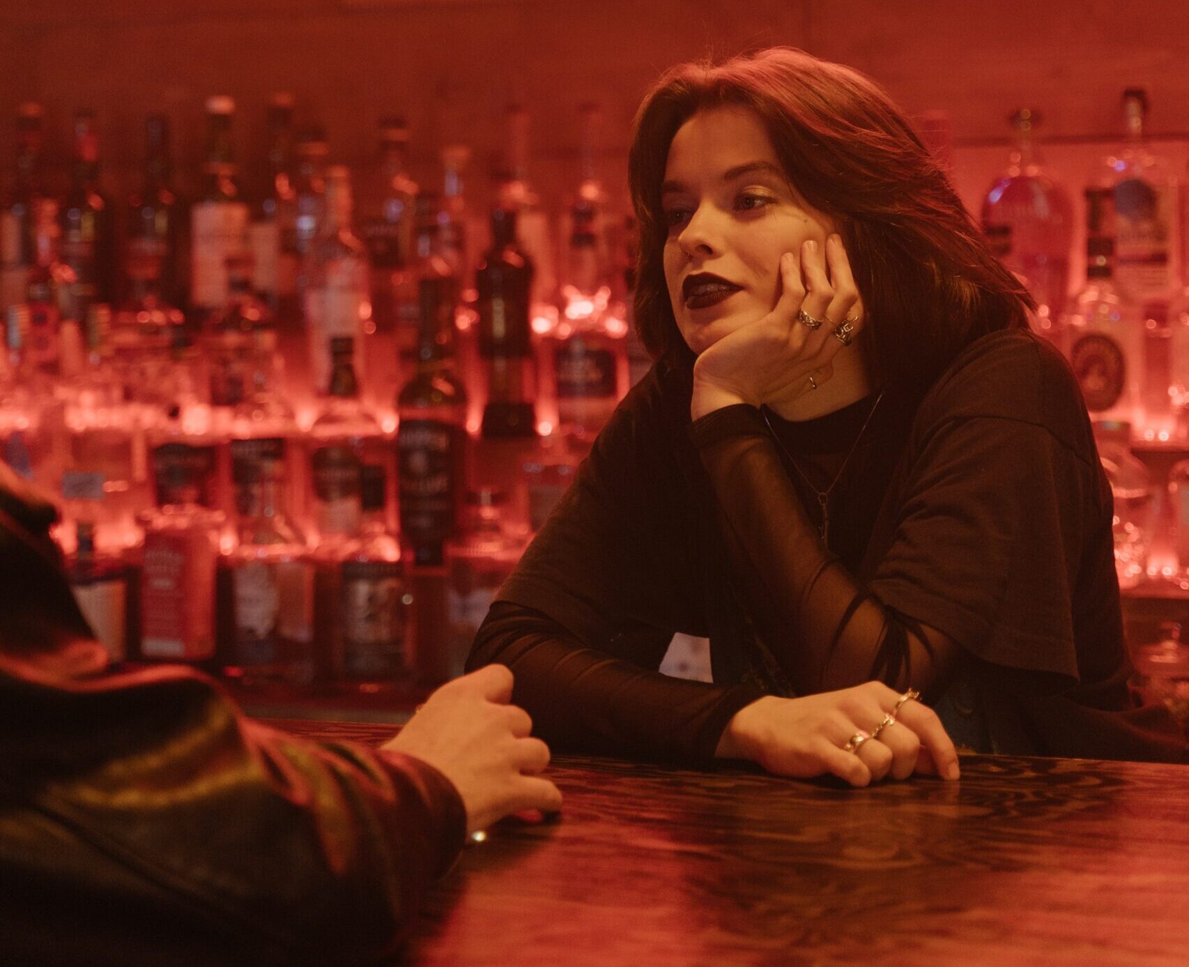 young femme person standing behind a bar with their hand on their chin, looking tired