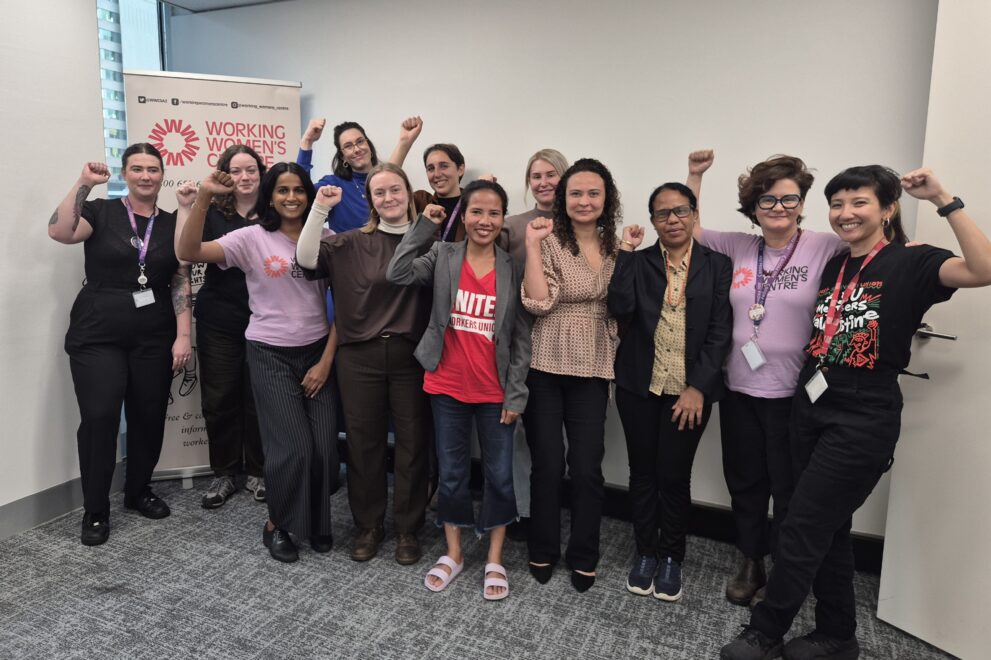 A group of women stand together in a room, smiling and raising their fists in solidarity. Behind them is a banner with the Working Women’s Centre logo.