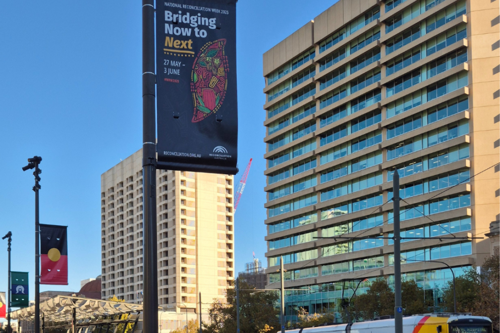 Street poles display banners for National Reconciliation Week 2025. One banner features the theme “Bridging Now to Next,” along with the dates 27 May – 3 June and a vibrant artwork. The Aboriginal and Torres Strait Islander flags are also visible on nearby banners.