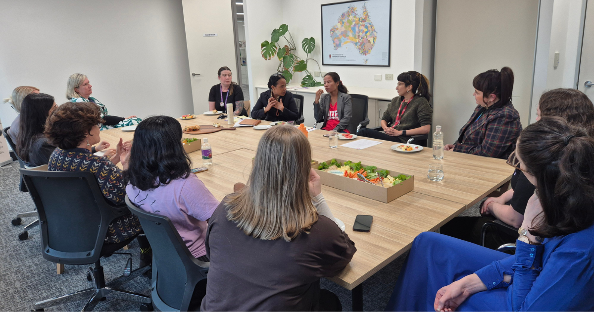 A group of women sit around a large meeting table engaged in conversation at the Working Women’s Centre South Australia. The table is set with lunch platters, water, and notepads. A large map of Australia hangs on the wall in the background.