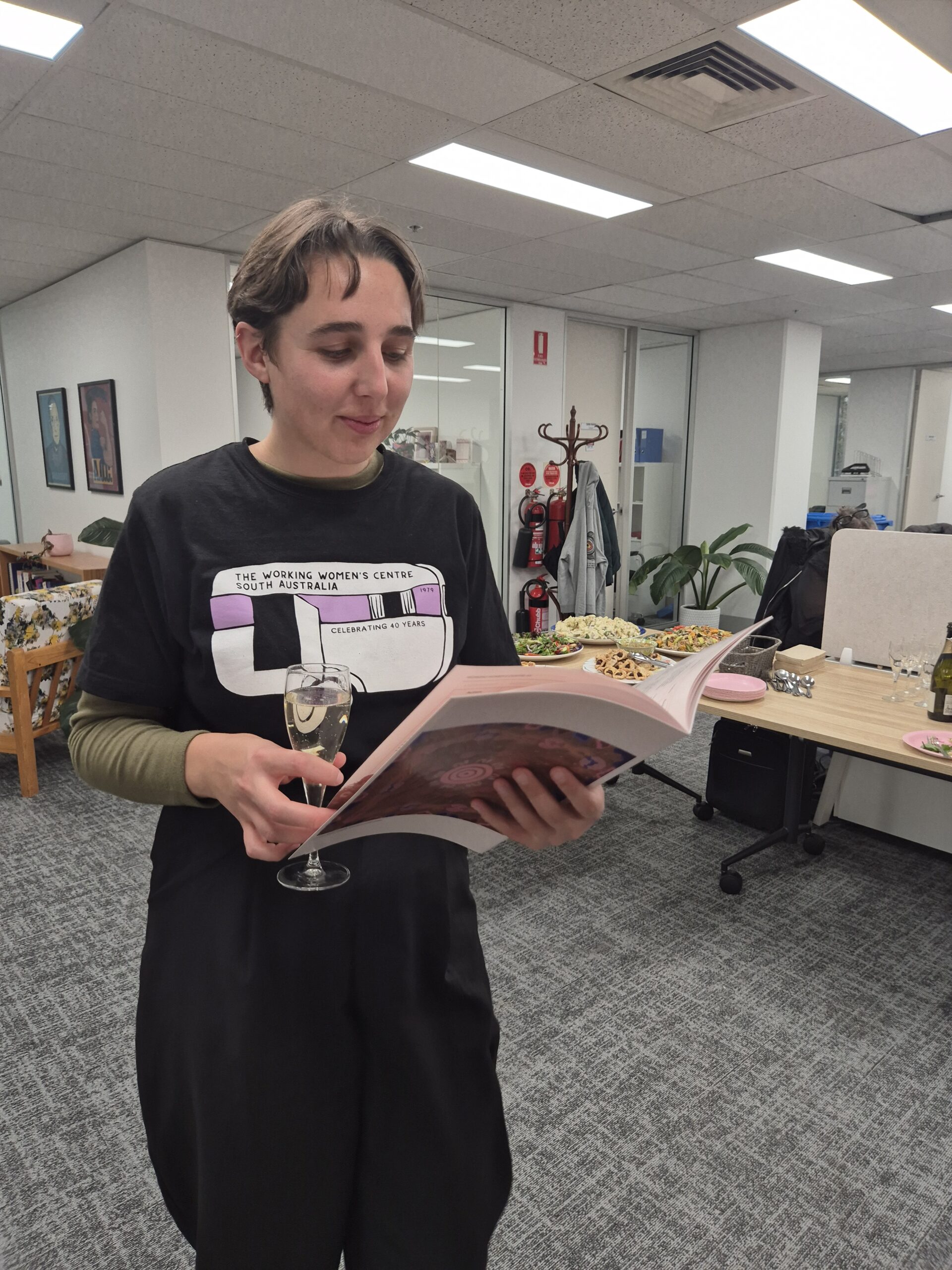 A person wearing a black "Working Women’s Centre South Australia" t-shirt holds a glass of sparkling wine and reads the Innovate Reconciliation Action Plan during the official launch event. In the background, an office space is set up with drinks, snacks, and an Aboriginal flag, marking the celebration of the RAP launch.