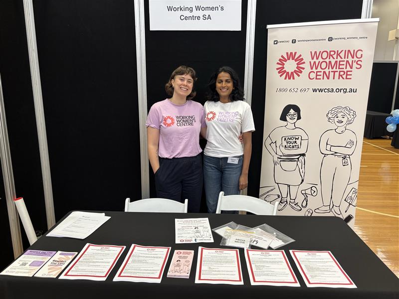 Two people standing and smiling at the Working Women’s Centre SA booth, with informational flyers and materials on the table, and a banner displaying the centre’s logo, contact details, and illustrated figures promoting workers' rights.