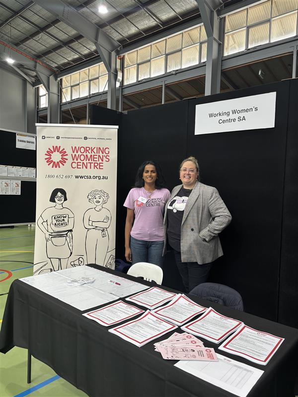 Two representatives standing at the Working Women’s Centre SA booth, smiling in front of a banner displaying the centre’s logo and worker rights illustration. The table is covered with informational flyers, forms, and pamphlets.