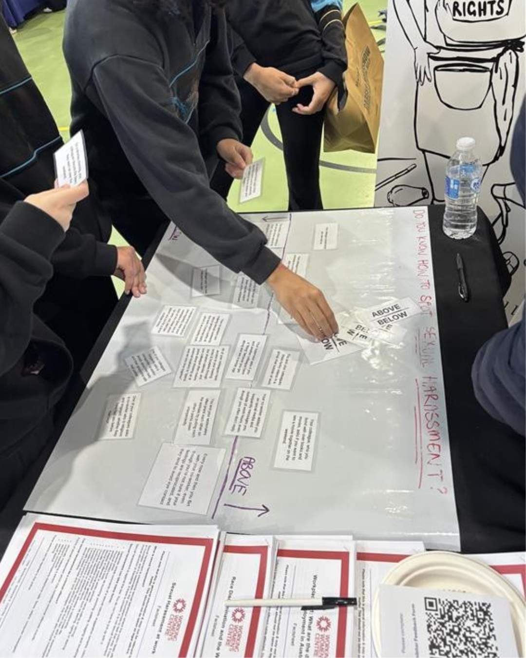 Several people participating in an interactive activity at a table, sorting cards with scenarios related to sexual harassment on a board marked 'Above' and 'Below the line.' The table also displays informational flyers and resources from the Working Women’s Centre SA."