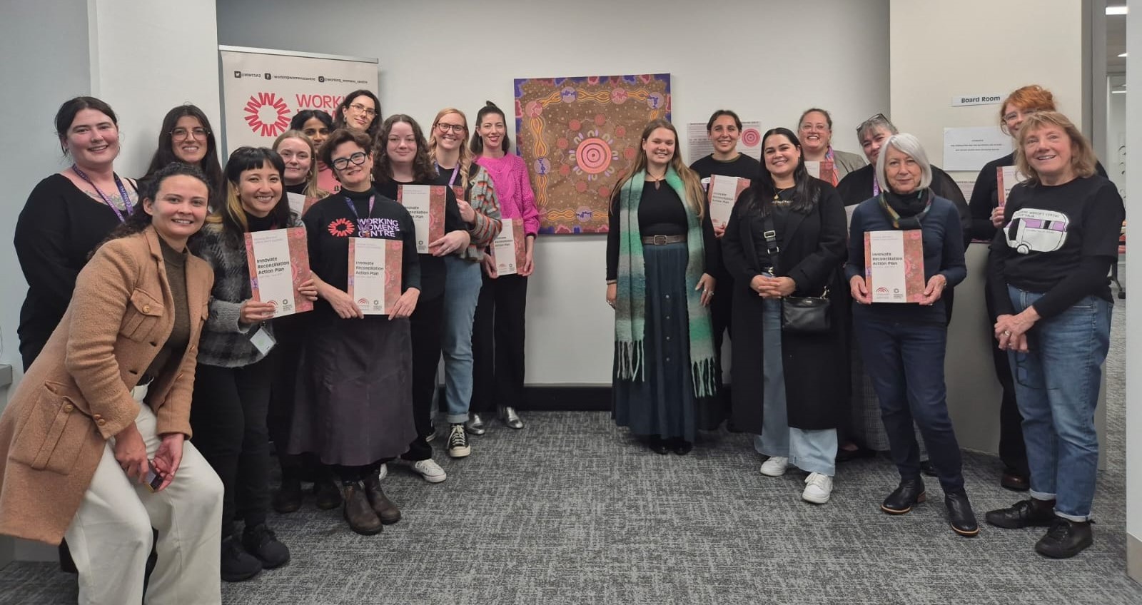 The Working Women’s Centre South Australia team stands together indoors, smiling and holding printed copies of a report. Behind them are a WWC SA banner and a vibrant piece of Aboriginal artwork.