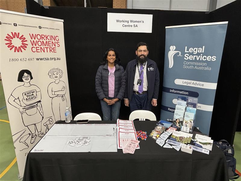 Two representatives standing behind a shared booth for the Working Women’s Centre SA and the Legal Services Commission of South Australia. The table displays brochures, flyers, and resources, with banners from both organisations in the background promoting legal advice and workers’ rights.