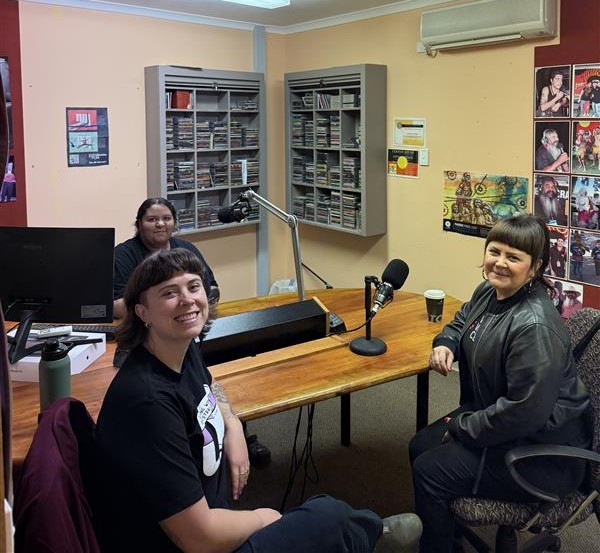 Three people sitting in a radio station studio, smiling at the camera. They are seated around a wooden desk with microphones, a computer, and audio equipment, with shelves of CDs and posters on the walls in the background.