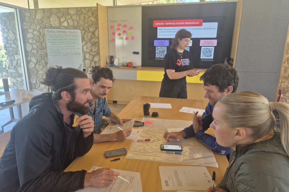 Small group of participants seated around a table during a workplace training session, discussing printed materials while a facilitator stands near a screen displaying presentation slides in the background.