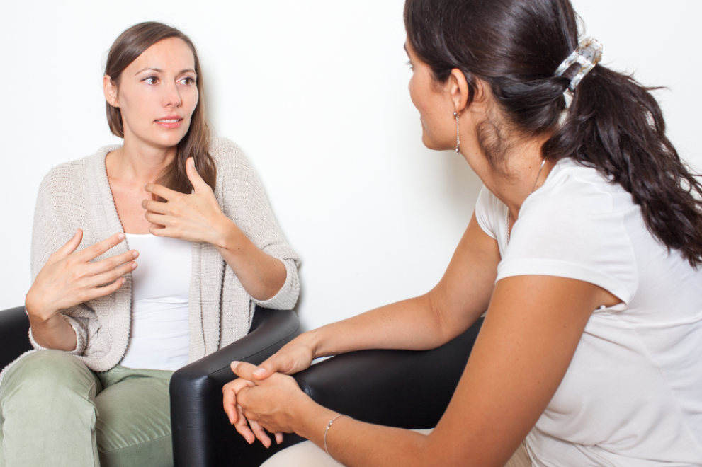 Two people seated and facing each other in a private setting, engaged in a supportive conversation. One person gestures while speaking, and the other listens attentively, suggesting a confidential discussion.