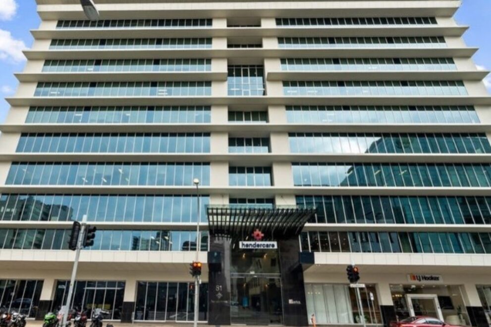 Modern multi-storey office building with a glass façade and white horizontal architectural features, viewed from street level at a city intersection. The building entrance is centred beneath a black canopy, with retail tenancies at ground level, traffic lights in the foreground, and a partly cloudy blue sky overhead.