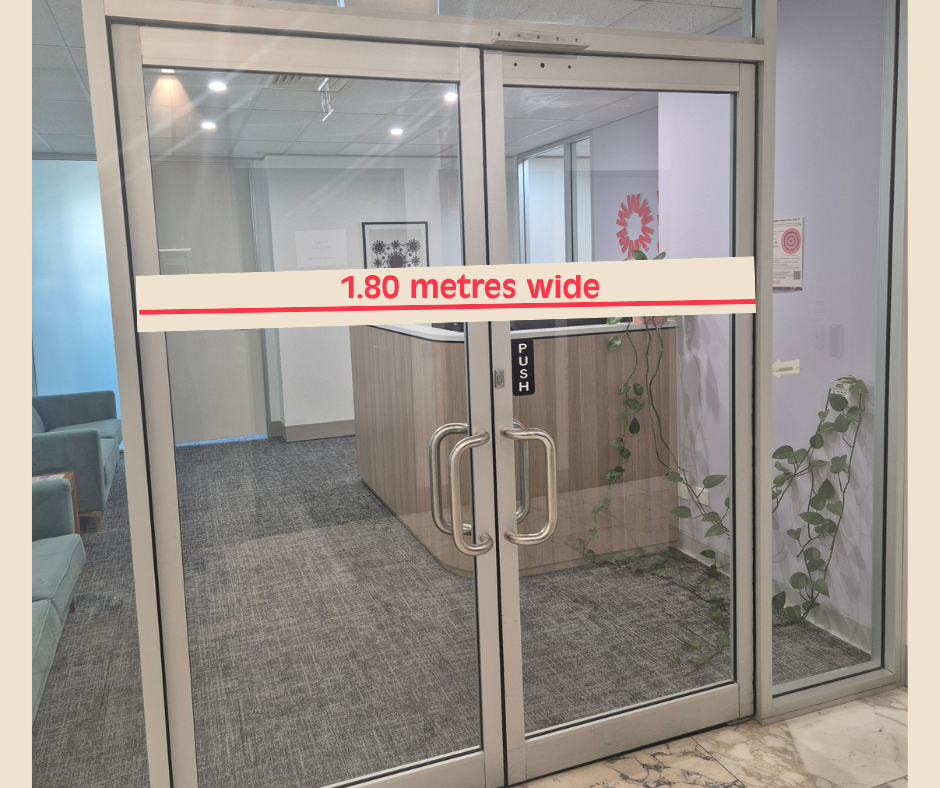 Glass double doors leading into a reception area, with a measurement label indicating the entrance is 1.80 metres wide. Beyond the doors is a reception desk, waiting area seating, framed wall artwork, and indoor plants. The space is well-lit, modern, and designed with a wide, accessible entryway.