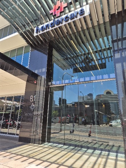 Close-up view of the entrance to the HenderCare building, featuring large glass doors, reflective windows, and a modern metal canopy overhead. The building number "81" is displayed beside the entrance, with reflections of the surrounding city streetscape visible in the glass façade.