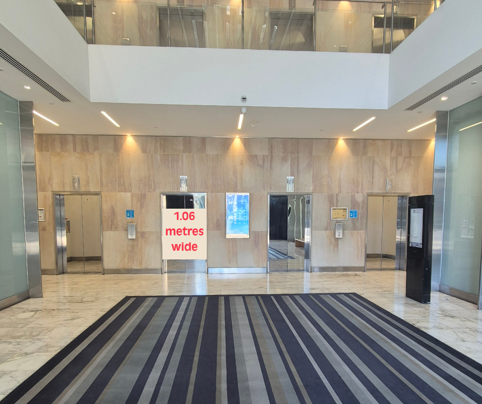 Office building lobby with four elevator doors set into a light stone wall. A large striped floor mat leads towards the lifts, and a sign displayed near one of the elevators indicates a width of 1.06 metres. The area is brightly lit with natural light from an upper-level atrium and overhead lighting, creating a clean and modern appearance.
