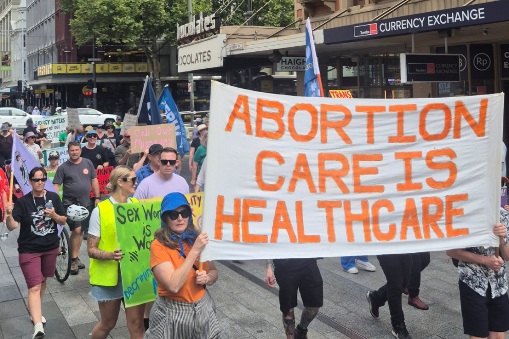 A large group of people march through Rundle Mall in Adelaide during a reproductive rights rally. At the front of the march, participants carry a large white banner with orange lettering that reads, “ABORTION CARE IS HEALTHCARE.” Protesters walk along the city street holding signs and flags, with shops, office buildings and pedestrians visible in the background. The atmosphere is peaceful and community-focused, with participants advocating for access to reproductive healthcare.