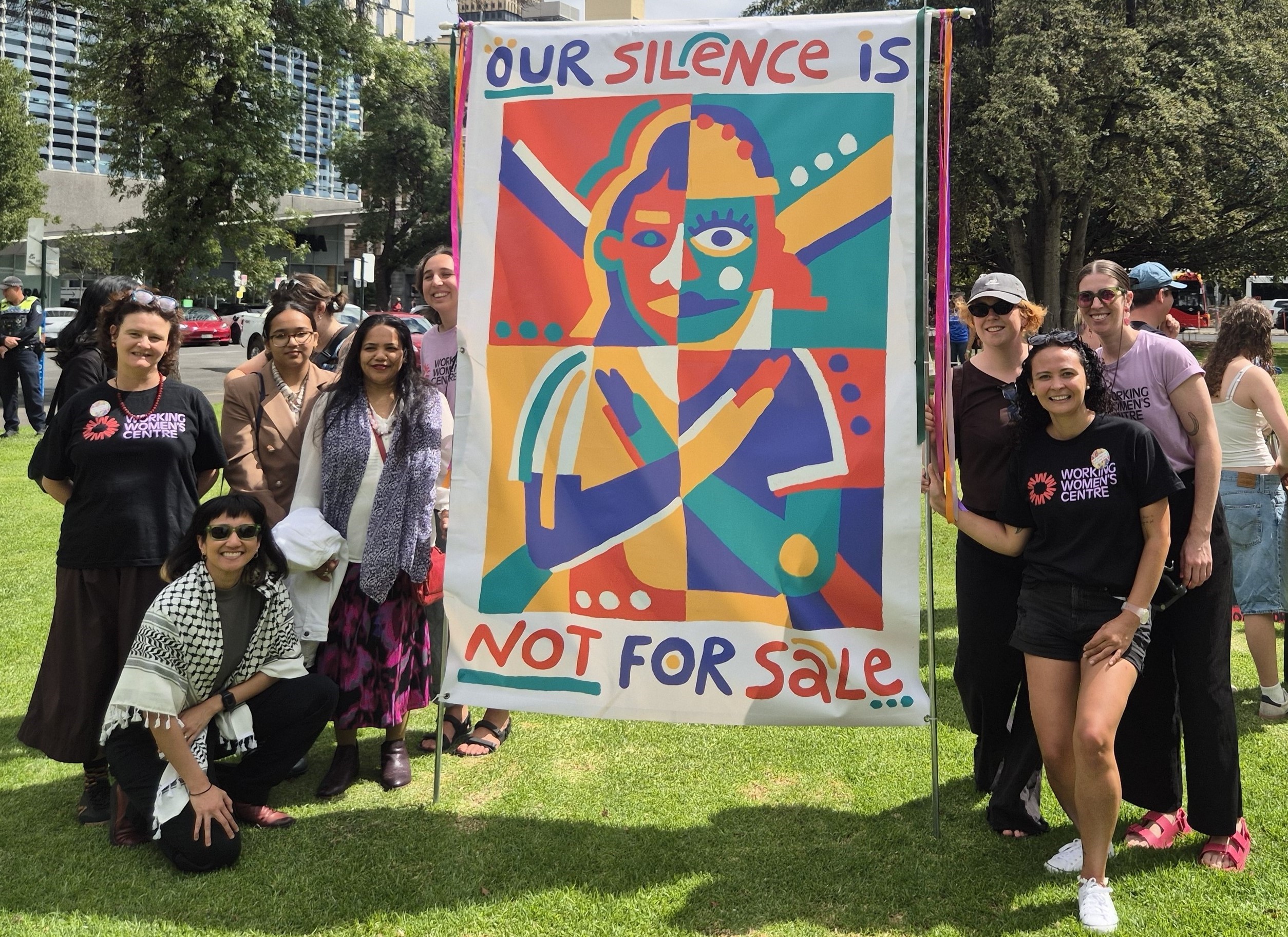 A group of Working Women's Centre staff and supporters stand in a park around a large, colourful banner reading 
