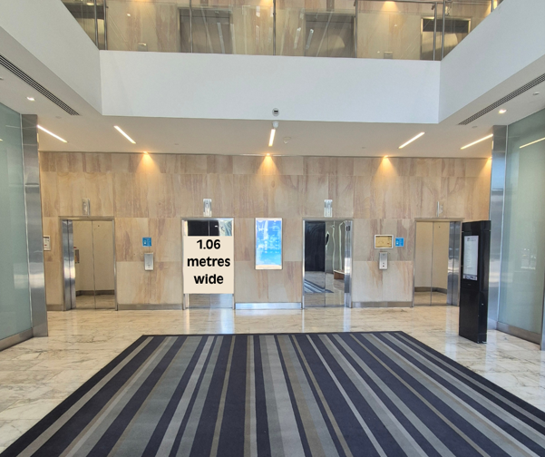 office building lobby with four elevator doors set into a light stone wall. A large striped floor mat leads towards the lifts, and a sign displayed near one of the elevators indicates a width of 1.06 metres. The area is brightly lit with natural light from an upper-level atrium and overhead lighting, creating a clean and modern appearance.