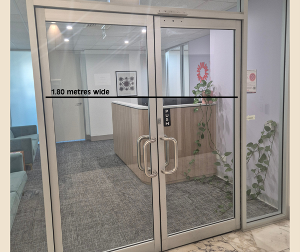 Glass double doors leading into a reception area, with a measurement label indicating the entrance is 1.80 metres wide. Beyond the doors is a reception desk, waiting area seating, framed wall artwork, and indoor plants. The space is well-lit, modern, and designed with a wide, accessible entryway.