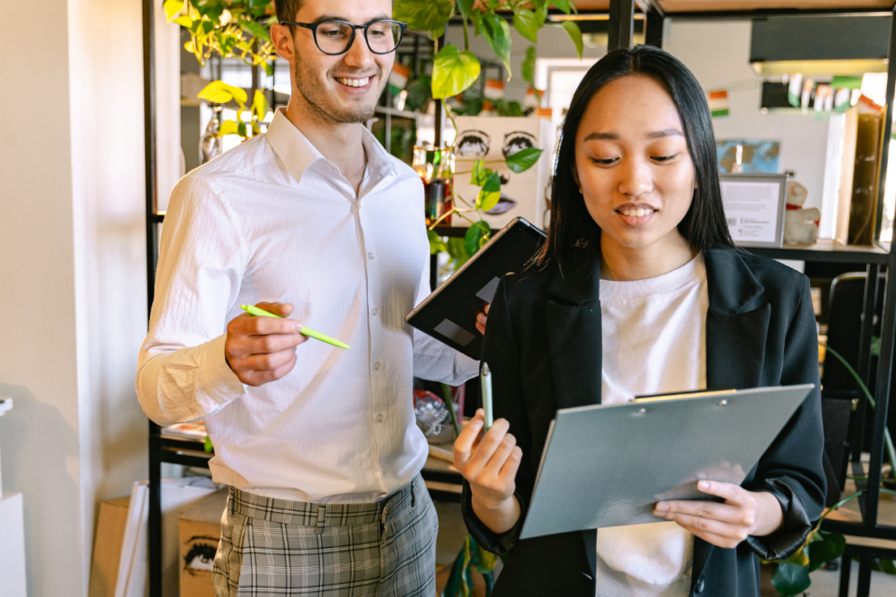 Two colleagues standing in an office, reviewing a clipboard together while discussing work. One person holds a tablet and pen, while the other gestures with a highlighter. Plants and shelving in the background create a bright, collaborative workspace.