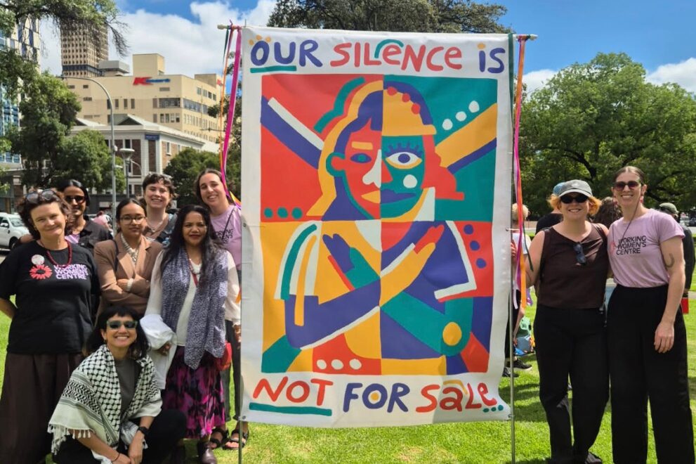 Group of women standing outdoors beside a colourful banner reading “Our Silence Is Not For Sale” at a public event.